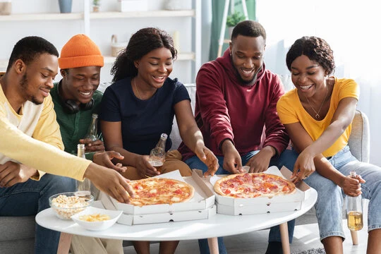 Five friends laughing as they reach for slices from two pizza boxes on a table